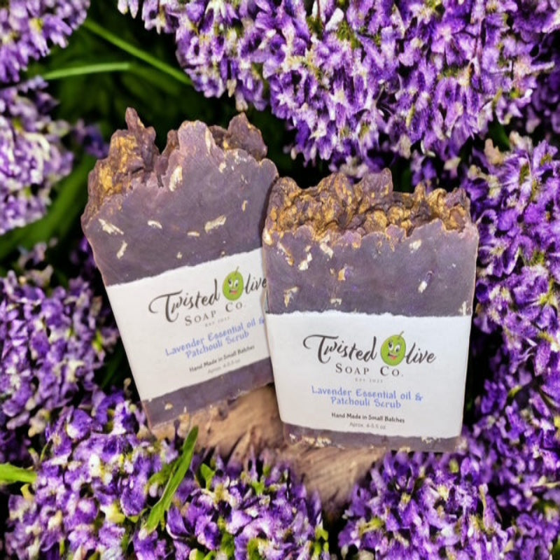 Two violet soap bars from Twisted Olive Soap Co. surrounded by lavender flowers. The soap has ingrained oatmeal as a loofah.