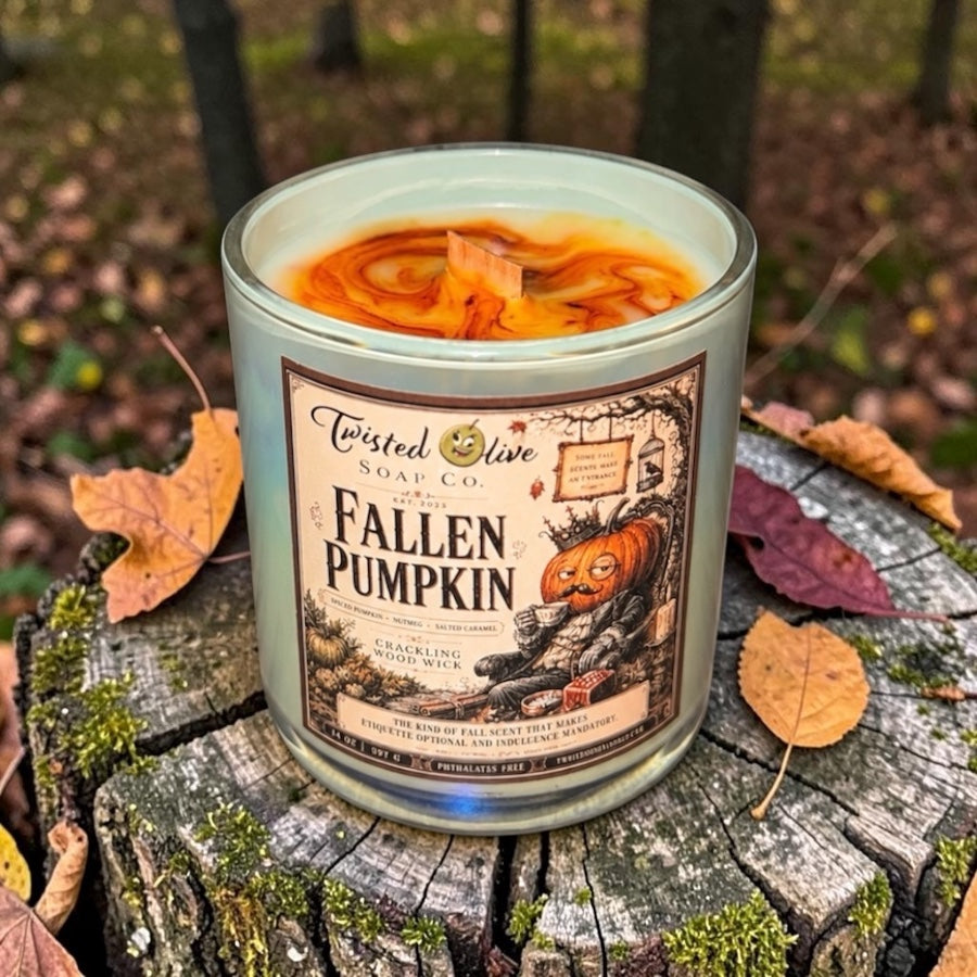 Fallen Pumpkin -Coconut Soy Wax Swirled on the surface with varations of orange colored wax kind of tye dyed Candle with a crackling wood wick in a sage colored irridescent glass candle jar. The Candle rests on an aged cut off tree trunk in the woods with scattered autumn leaves. The Label is antique sketched of a fallen pumpkin head man leaning against a tree trunk
