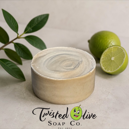 A round Tallow olive oil soap round soap bar with a swirled cream and soap ash top, styled with green leaves and limes on a neutral background. The Twisted Olive Soap Co.