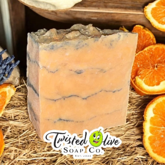 Bar of soap that is a light orangey color with thin bands of charcoal running through with orange slices and dried flowers on a bed of straw featuring Twisted Olive Soap Co logo.