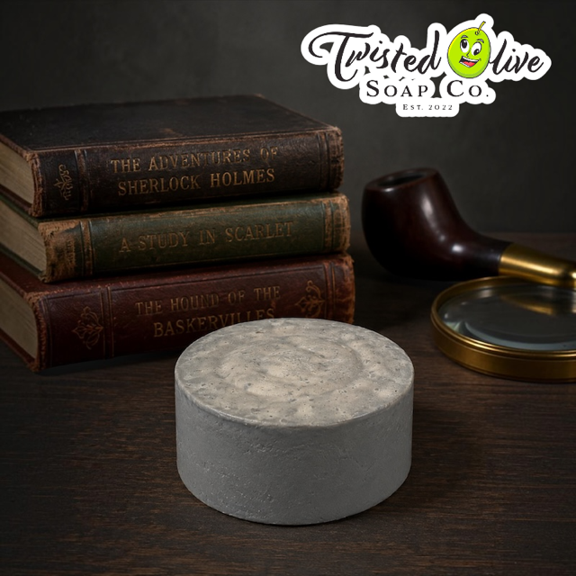 Twisted Olive Soap Co Est. 2022 at top right of picture. Grey round soap sits on a brown surface with a Three antique books piled on top of each other with an dark wood tobacco pipe with a brass tip and brass small tray next to the pipe.   