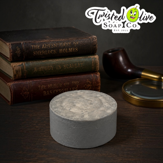 Twisted Olive Soap Co Est. 2022 at top right of picture. Grey round soap sits on a brown surface with a Three antique books piled on top of each other with an dark wood tobacco pipe with a brass tip and brass small tray next to the pipe.   