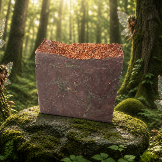 A rectangular Brown soap with mossy green veins scrolling through sitting on a mossy rock in the middle of a green forest with the sun shining through the tall trees and  little fairies peaking out from an ancient tree on the right and one peaking out on the left.  Moss covers most of the forest ground