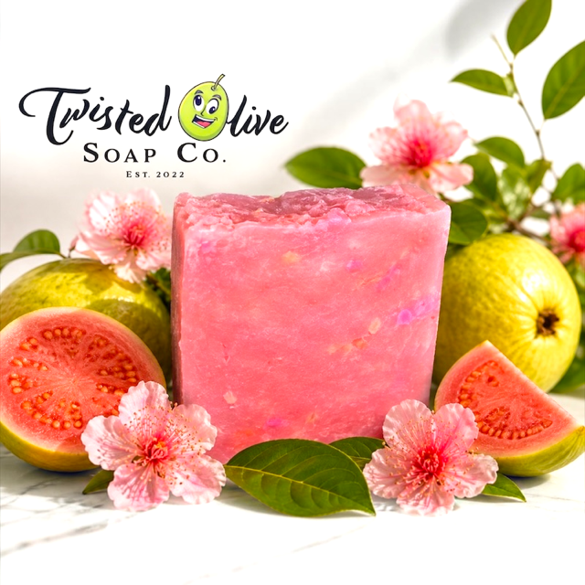 A rectangular bar of pink soap with some white spots from Twisted Olive Soap Co. is displayed in a styled arrangement with sliced guava, pink flowers, and green leaves on a white marble surface. The Twisted Olive Soap Co. logo is at the top left
