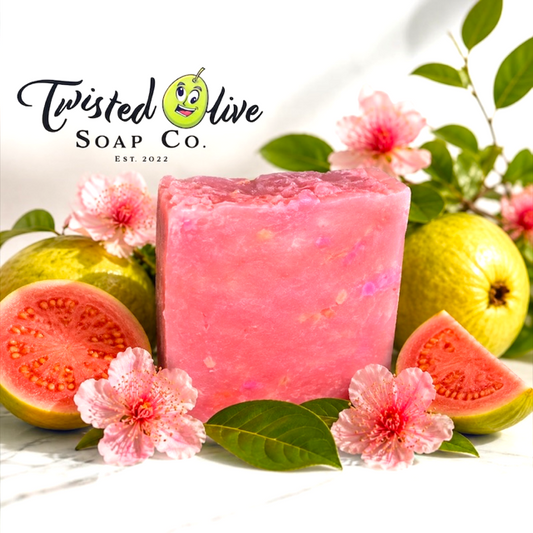 A rectangular bar of pink soap with some white spots from Twisted Olive Soap Co. is displayed in a styled arrangement with sliced guava, pink flowers, and green leaves on a white marble surface. The Twisted Olive Soap Co. logo is at the top left
