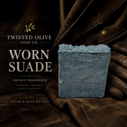 A single rectangular bar of soap labeled Twisted Olive Soap Co. 'Worn Suade' stands on dark brown fabric beside a brown leather jacket and gloves.Unisex Fragrance. Cypress· Woodsy·Black Currant.  Tallow and Olive oil Soap