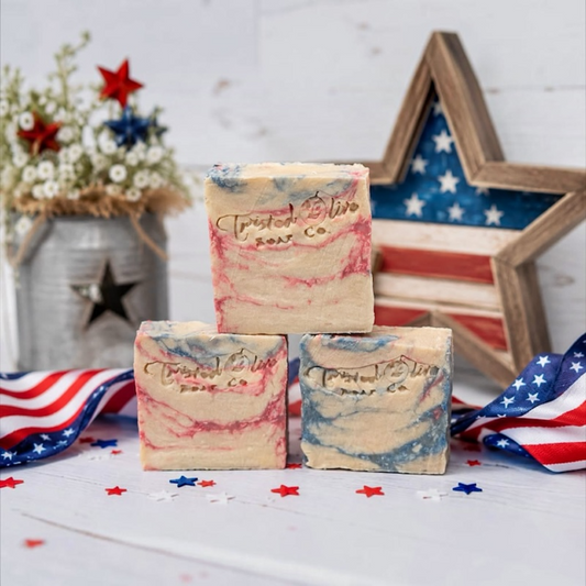 Three handmade patriotic soap bars by Twisted Olive Soap  Co. displayed witha n American Flag ribbon, rustic wooden flag star, and rid white and blue decorations on a white background sitting on a white cloth with star confetti